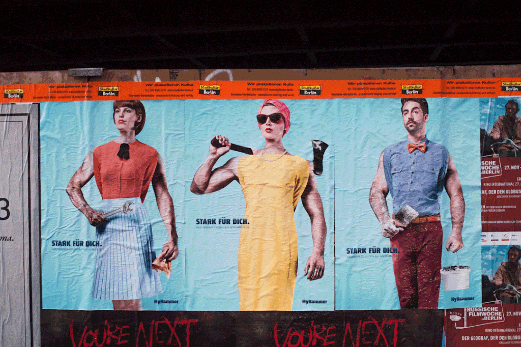 Bauzaun-Plakate mit Personen, hinter denen die Arme von Profis mit Werkzeug hervorschauen